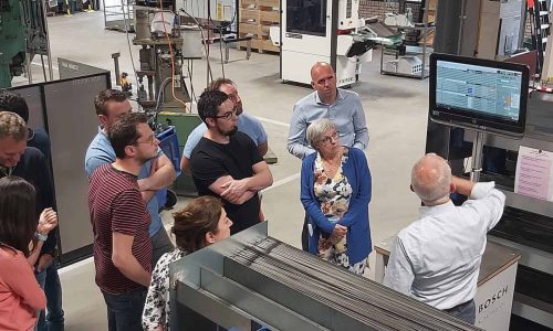 Kunststof scharnieren demonstratie in fabriek, mensen luisteren aandachtig naar instructie, technologische productieomgeving, Bosch Scharnieren expertise.